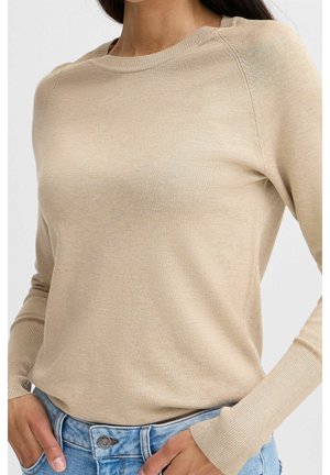 Kobieta ubrana w beżowy, długi sweter z prążkowanymi mankietami, wsunięty w jasnoniebieskie dżinsy, z rękami spoczywającymi blisko kieszeni.