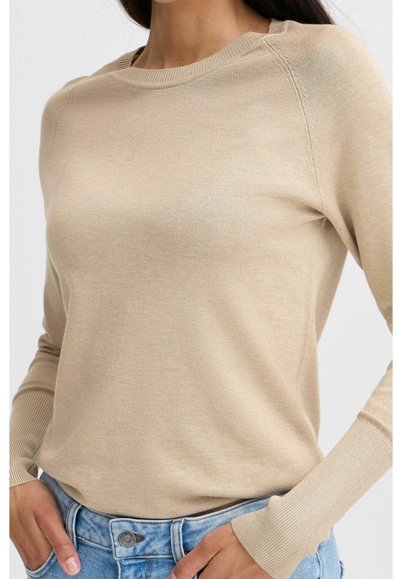 Femme portant un pull beige à manches longues avec des poignets côtelés, rentrée dans un jean bleu clair, les mains reposant près des poches.