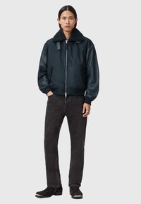 Laivaston sininen pommikoneen takki, jossa on teksturoitu tummanvihreä shearling-kaulus, vetoketjusuljenta ja kaksi sivutaskua, yhdistettynä mustiin farkkuihin ja mustiin saappaisiin.