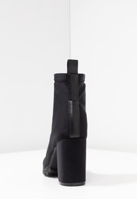 Bottine noire à talons hauts fabriquée en tissu avec un accent en cuir verni à l'arrière et un large talon. Texture lisse avec des détails de couture minimaux.