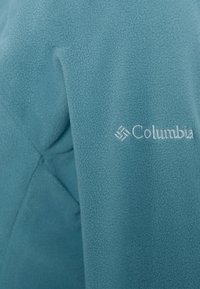 Petrolowa kurtka z polaru o gładkiej fakturze, z haftowanym logo "Columbia" w białym kolorze. Dopasowany krój z subtelnymi detalami przeszyć.