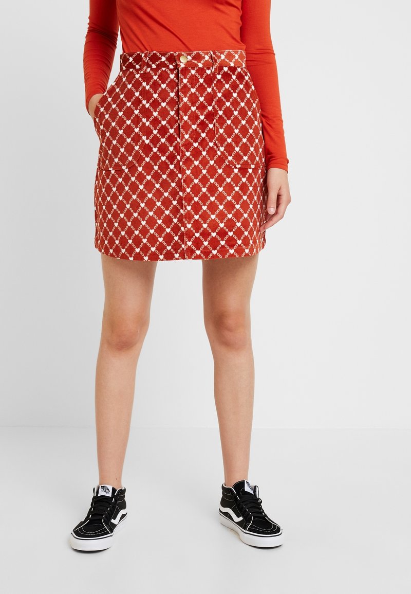 Femme portant une jupe rouge à motifs avec des boutons sur le devant et des poches, un haut orange à manches longues, et des baskets montantes noires avec semelles blanches.