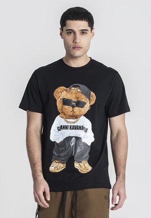 Sort t-shirt med et stort grafisk tryk af en bjørn i solbriller og en hvid skjorte med teksten "GIANNI KAVANAGH," parret med grå bukser og guld sko.