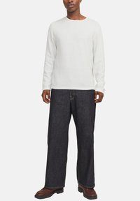 Eine Person trägt ein strukturiertes weißes Langarmshirt, lockere dunkelblaue Jeans und braune Lederschuhe und steht vor einem einfarbigen Hintergrund.