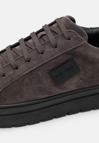 Baskets en daim gris avec une surface texturée, dotées de lacets ronds, d'une semelle en caoutchouc noire et d'un détail logo rectangulaire sur le côté.