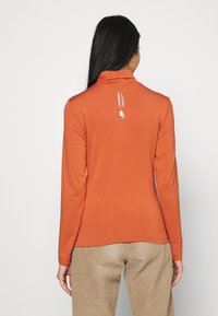 Kvinna med långt svart hår som bär en orange långärmad Nike sporttröja och beige byxor, sedd bakifrån.