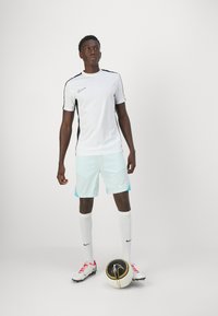 Conjunto de fútbol de Nike que incluye una camiseta blanca de manga corta con acentos negros, pantalones cortos azul claro, calcetas blancas hasta la rodilla y botas negras sosteniendo un balón.