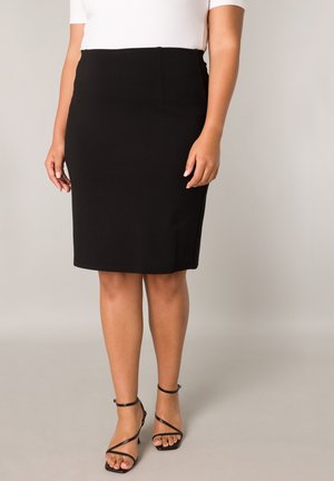 Vrouw die een zwarte kokerrok tot op de knie, een wit bovenstuk en zwarte sandalen met hoge hakken en bandjes draagt, staand tegen een neutrale achtergrond.