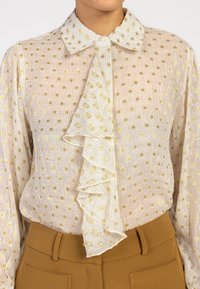 Sheer, patterned blouse in pale cream with gold dots, featuring a ruffled front and a collared neckline. Paired with brown high-waisted pants.