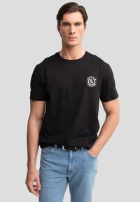 Schwarzes kurzärmliges Baumwoll-T-Shirt mit weißem rundem Logo auf der linken Brust, kombiniert mit hellblauen Jeans und einem schwarzen Gürtel.