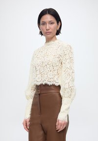 Γυναίκα με κοντά σκούρα μαλλιά που φοράει εκρού μπλούζα με δαντέλα και μακρύ μανίκι και καφέ δερμάτινο παντελόνι με ψηλή μέση, κοιτάζοντας προς τα εμπρός, σε λευκό φόντο.