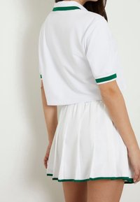 Weißes Kurzarm-Polo mit grünen Akzenten, kombiniert mit einem weißen plissierten Rock, der grüne Verzierungen am Saum hat. Glatte Stofftextur.