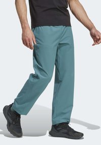 Türkise, leichte, technische Stoffhosen mit lockerer Passform, seitlichen Taschen und glatter Textur. Tragen mit schwarzen Sportschuhen.