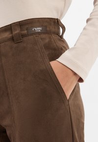 Close-up van een hand in de zak van bruine suède broek met een "CMRV Future" label, gecombineerd met een lichtbeige blouse met lange mouwen.