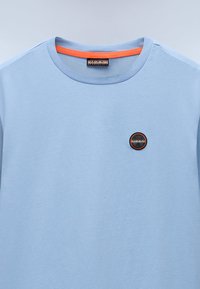 Světle modré bavlněné tričko s kulatým výstřihem, se dvěma oranžovými detaily uvnitř límečku a černobílým logem s oranžovým písmem na přední straně.