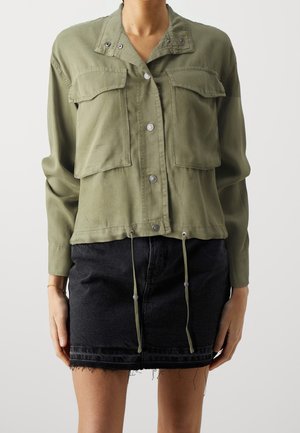 Vrouw draagt een olijfgroene jasje met knopen, grote voorzakken en trekkoorden, gecombineerd met een zwarte, rafelige spijkerbroek mini rokje.