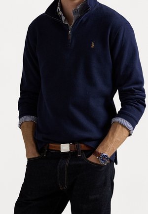 Homme portant un pull marine à fermeture éclair quart, par-dessus une chemise rayée à col, un jean foncé, une ceinture marron et une montre-bracelet avec un cadran bleu et un bracelet en cuir marron.