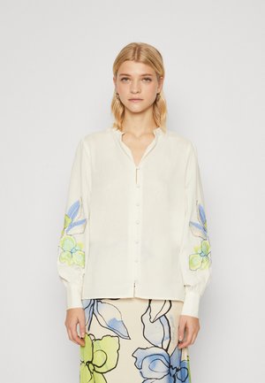 Crème kleurige blouse met een kraaghalslijn, lange mouwen met blauwe en groene geborduurde bloemen, en een knoopsluiting aan de voorkant.