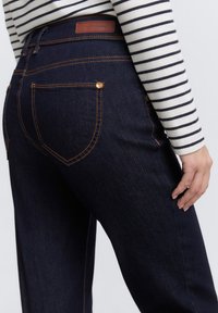 Mørkeblå denimjeans med høy midje, oransjefargede sømmer og en lærmerke bak. Har to baklommer og en rett passform.