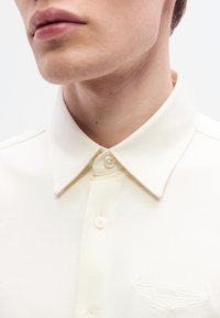 Πόλο πουκάμισο σε κρεμ χρώμα με κλασικό γιακά, δύο κουμπιά και κεντημένο λογότυπο στην αριστερή πλευρά. Ομαλή υφή υφάσματος.