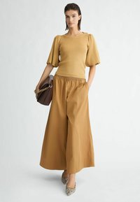 Top a maglia con maniche a sbuffo di un giallo dorato, abbinato a pantaloni larghi a vita alta in un marrone terroso e décolleté a punta con fantasia.