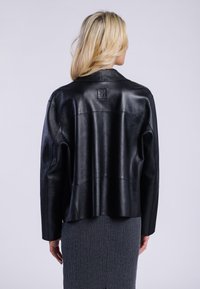 Giubbotto di pelle nera con un fit rilassato, caratterizzato da un design cucito e una patch con logo sul retro, indossato sopra una gonna grigia testurizzata.