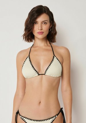 Femme portant un bikini triangle texturé crème avec bordure noire et détails à nouer, debout devant un fond clair uni.