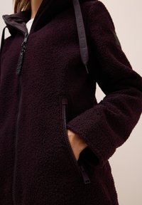 Veste en polaire de couleur violet foncé, avec fermeture éclair, poches zippées et capuche ajustable en tissu texturé.