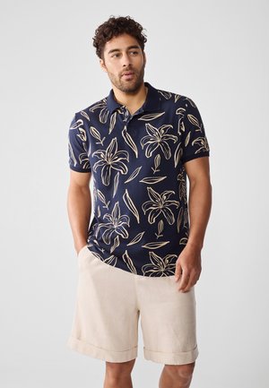 Polo shirt marine avec un motif floral crème, à manches courtes et col, associé à un short en lin beige, offrant une coupe décontractée.