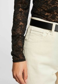 Top in pizzo nero con motivo floreale e maniche lunghe, abbinato a pantaloni di denim chiaro e una larga cintura in pelle nera.