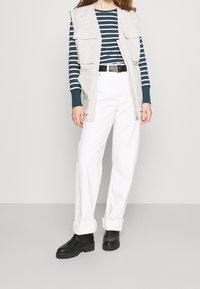 Ljusgrå ärmlös väst med två framfickor, marinblå och vit randig långärmad skjorta, vita vida byxor och svarta ankelboots.