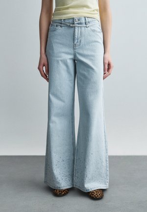 Osoba nosi svetlomodré džínsové nohavice širokého strihu s dekoratívnymi detailmi pri leme, svetložlté tričko a topánky so vzorom leopardej kože.