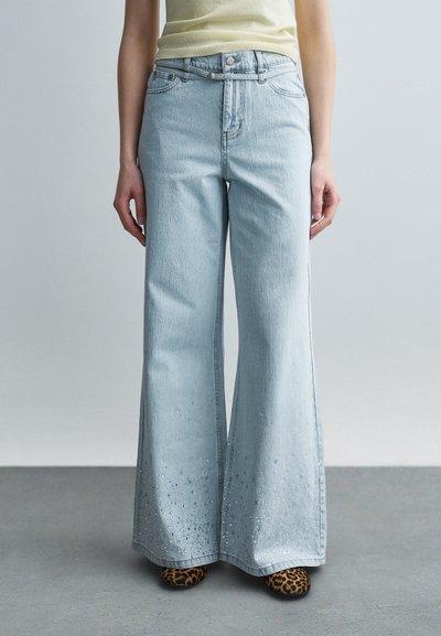 Personne portant un jean large bleu clair avec des détails décoratifs près de l'ourlet, un haut jaune clair et des chaussures imprimé léopard.