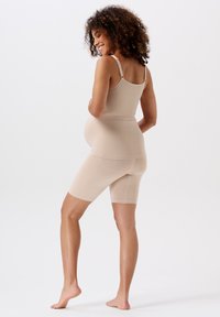 Zwangere vrouw met krullend haar in een beige tanktop en shorts staat blootsvoets, glimlachend en kijkt over haar schouder tegen een effen achtergrond.