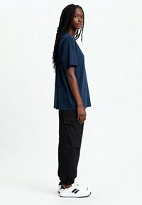 Jūras spēku īsām piedurknēm T-krekls apvienots ar melnām cargo biksēm, kas aprīkotas ar sānu kabatām un elastīgām aprocēm. Valkāts ar baltām sporta kedām.