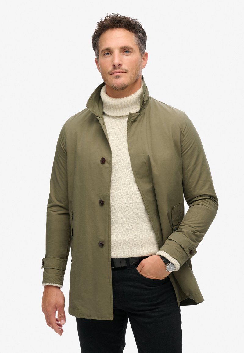 Manteau léger vert olive avec col montant, boutons marron et poches latérales, superposé sur un pull à col roulé crème et un pantalon noir.