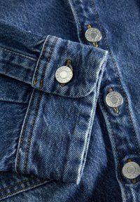 Niebieska dżinsowa tkanina z zbliżeniem na kołnierz koszuli i srebrne guziki. Szycie jest widoczne w pomarańczowej nitce, podkreślające design.