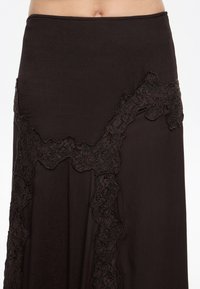 Środkowa część ciała ubrana w ciemnobrązową spódnicę z kwiatową koronkową lamówką tworzącą abstrakcyjny wzór na tkaninie.