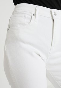 Jeans de denim blanco con una cintura alta, bolsillos frontales, costuras sutiles y cierre de botón metálico, que exhiben una tela texturizada.