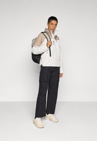 Hvid og beige jakke med lynlås og høj krave, parret med sorte cargo bukser og beige sneakers. Modellen bærer en sort rygsæk.