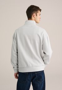 Hanorac tip pulover gri deschis cu guler înalt, manșete și tiv striate, asortat cu blugi albastru închis. Material neted, croială lejeră.