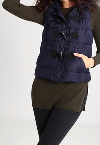 Marinblått pufferväst med knappar och toggle-stängningar, med quiltad textur och framfickor, bärs över en ribbad mörkgrön topp.