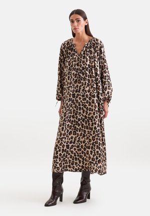 Kvinde iført lang leopardprint kjole med lange ærmer og sorte støvler med hæl, stående mod en ensfarvet lys baggrund.