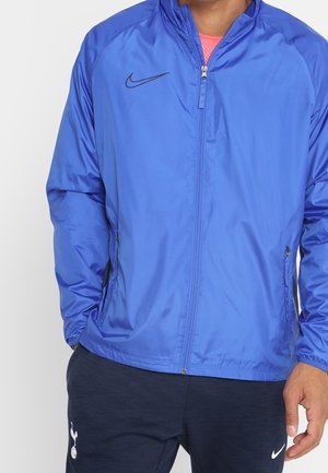 Mężczyzna w niebieskiej kurtce Nike na zamek błyskawiczny, różowej koszulce i ciemnoniebieskich spodniach Nike z logo Tottenham Hotspur.