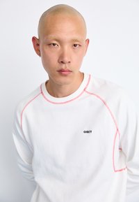 White sweatshirt with contrast red stitching, featuring a small black "OBEY" logo on the chest. Smooth texture, crew neck design.