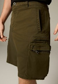 Shorts cargo verts olive avec une poche zippée et des rabats latéraux, fabriqués en tissu lisse. Dotés de passants de ceinture et de coutures renforcées.