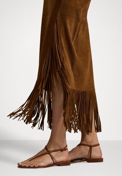 Noge v rjavi antilop suknji z dolgimi resicami in ravnimi svetlo rjavimi usnjenimi sandali z zankami okoli gležnjev na enobarvnem ozadju.