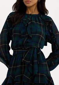Grønn og svart rutete bluse med rynket halslinning, lange ermer og et belte i midjen. Mykt stoff med en teksturert, lagdelt utseende.