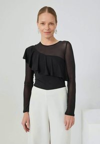 Schwarzes Langarmoberteil aus durchsichtigem Material mit Rüschendesign auf der Vorderseite. Trägt weiße, weit geschnittene Hosen, um das Outfit zu präsentieren.