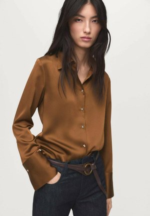 Femme aux longs cheveux bruns portant un chemisier en satin marron à boutons, rentré dans un jean foncé avec une ceinture marron, se tenant devant un fond uni.